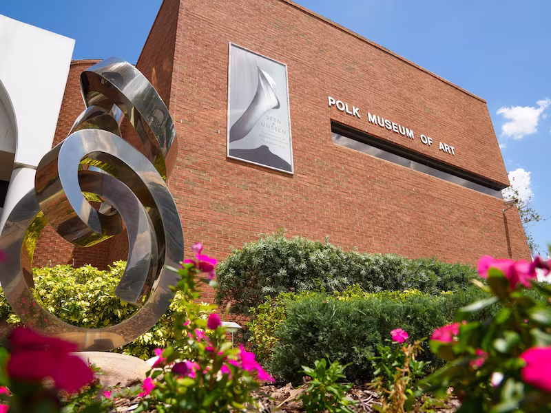 Exterior of Polk Museum of Art building with metal sculpture and pink flowers in frame.