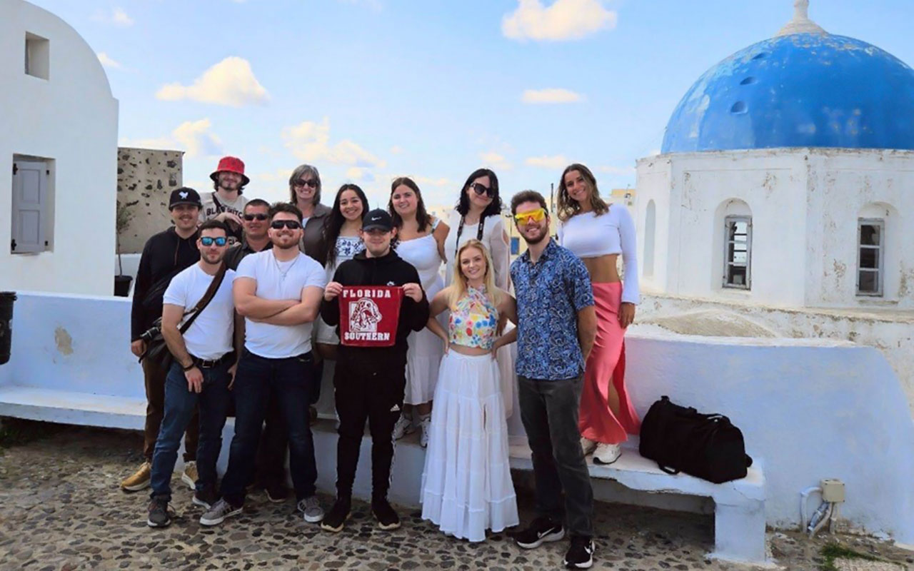 Florida Southern students with accompanying faculty posing together for a group photo with Florida Southern College Moccasins flag on an Italy trip with a waterside city landscape in the background. 
