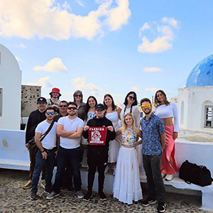 Florida Southern students with accompanying faculty posing together for a group photo with Florida Southern College Moccasins flag on an Italy trip with a waterside city landscape in the background. 